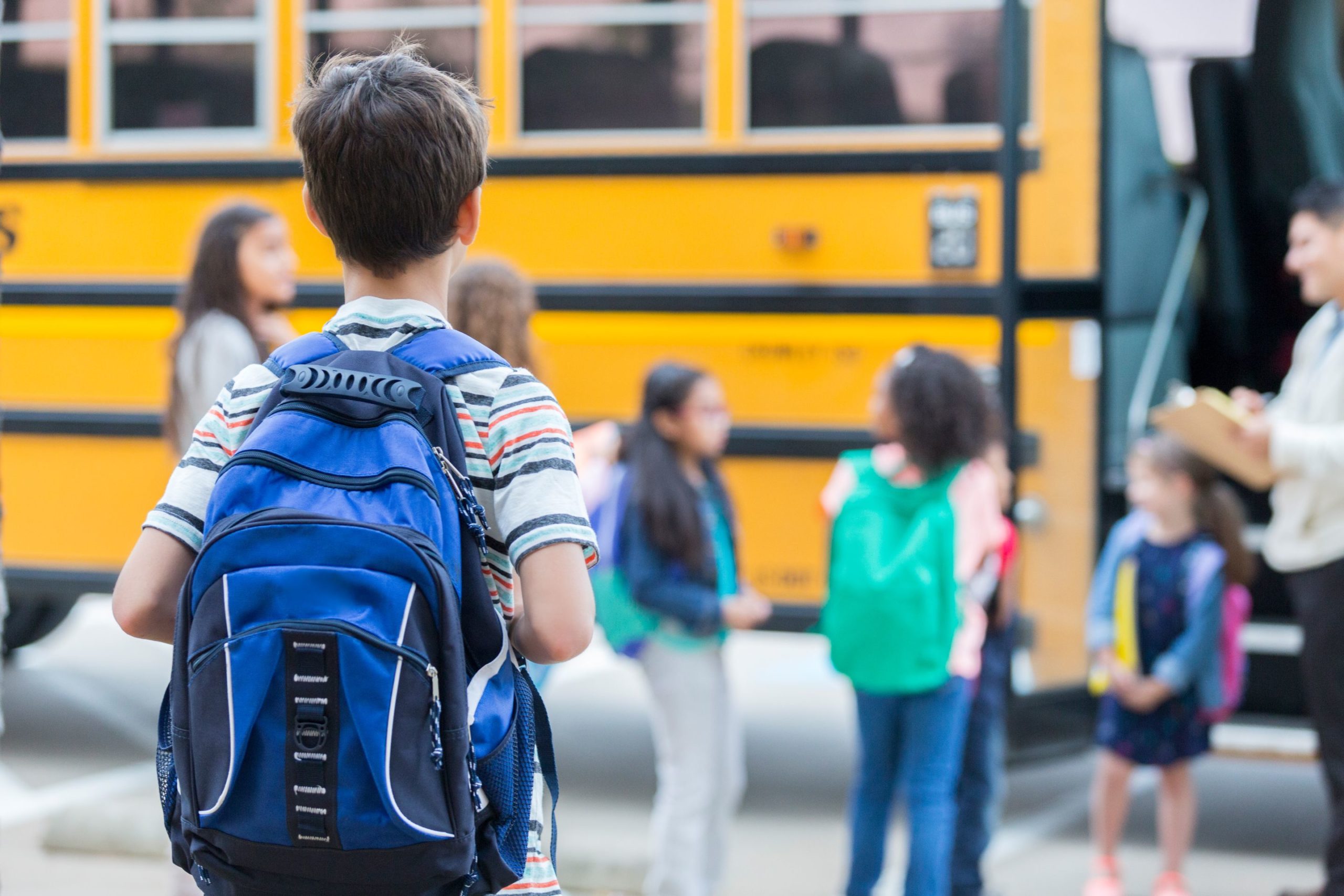 Rear view of elementary age boy waiting to get on school bus. His classmates are loading the bus in the background.